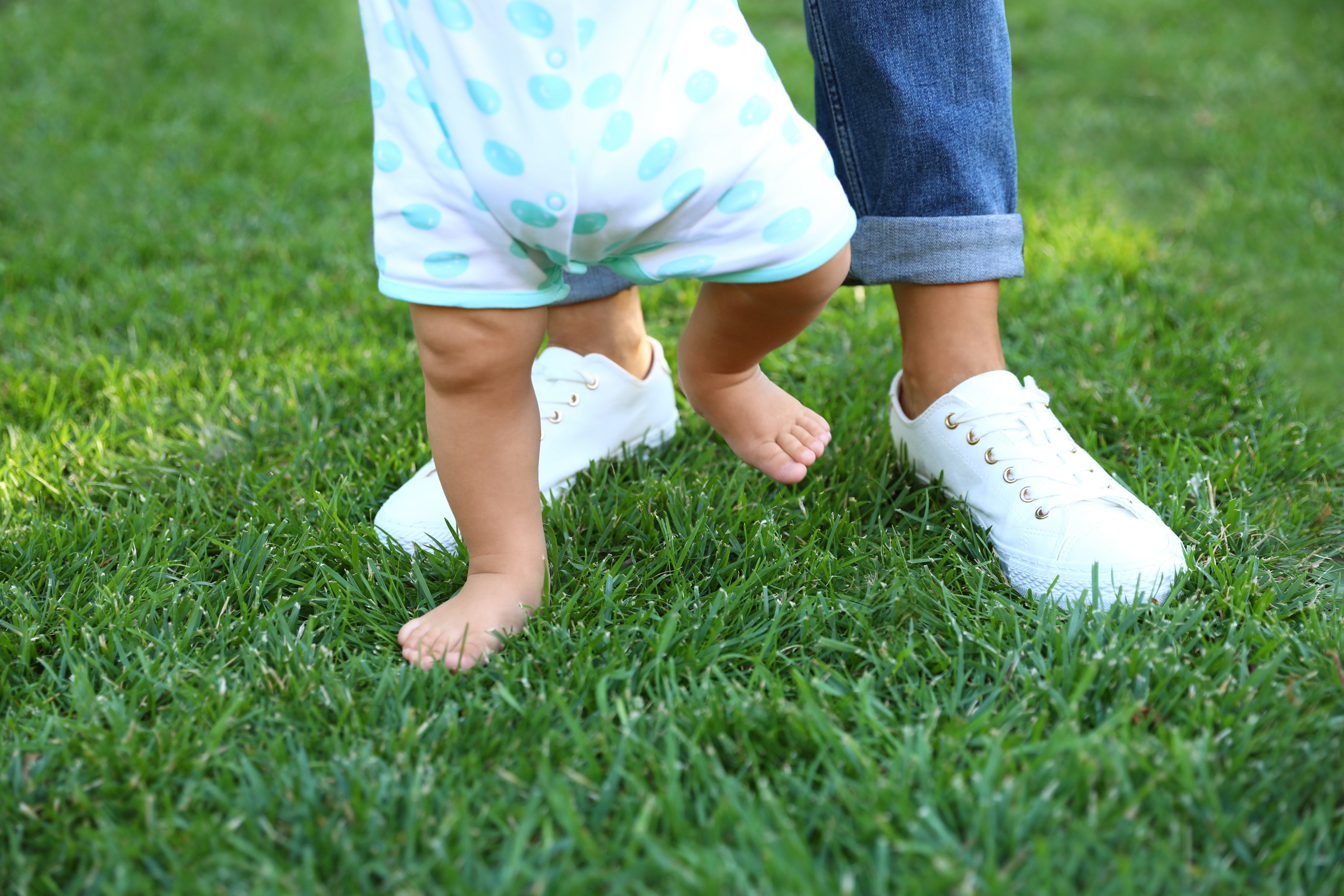 Cute Little Baby Learning to Walk with His Nanny on Green Grass Outdoors, Closeup